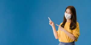 young-asia-girl-wear-medical-face-mask-shows-something-blank-space-with-dressed-casual-cloth-look-camera-social-distancing-quarantine-corona-virus-panoramic-banner-blue-background